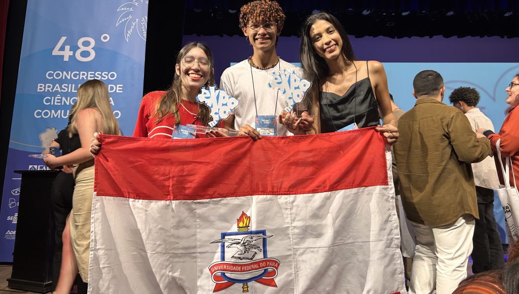 Fotografia posada das estudantes Gabriela, Luiza e Marcos Maia. Eles seguram um troféu em formato do mapa do Brasil e a bandeira da UFPA.