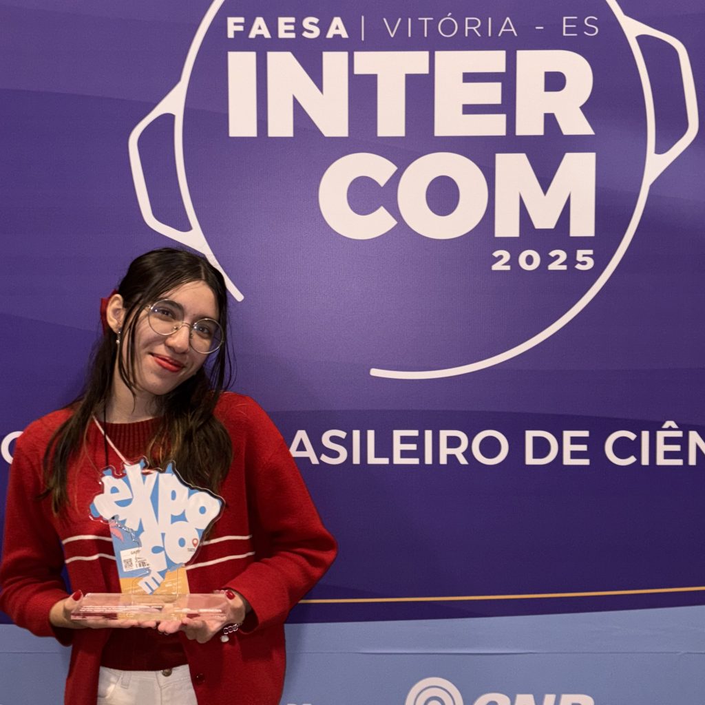Fotografia posada da estudante Gabriella Daud. Ela está a frente de um banner escrito "intercom 2025" e segura um troféu no formato do mapa do Brasil.