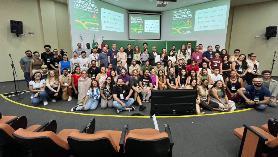 Fotografia mostra grupo de pessoas em pé posando para a foto. Ao centro estão o reitor e a vice-reitora da UFPA segurando uma folha de papel. Atrás do grupo vê-se três telas de projeção.