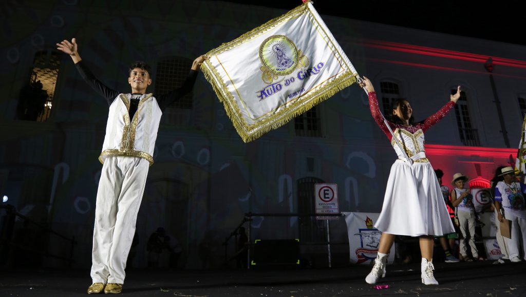 Fotografia de um casal de mestre sala e porta bandeira. Eles estão vestindo roupas brancas com detalhes dourados e exibem uma bandeira também branco, com detalhes dourado, com o símbolo do Auto do Círio.
