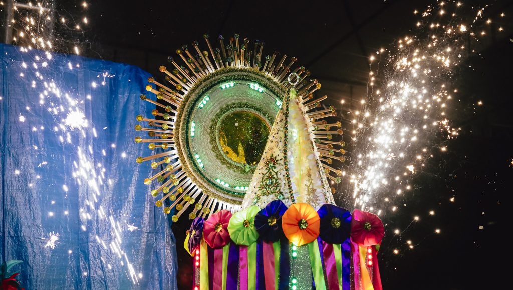 Fotografia registra um manto sobre uma estrutura com diversas fitas coloridas penduradas. Ao fundo, é possível ver um arco brilhante rodeado de luzes. No céu, luzes de fogos de artifício.