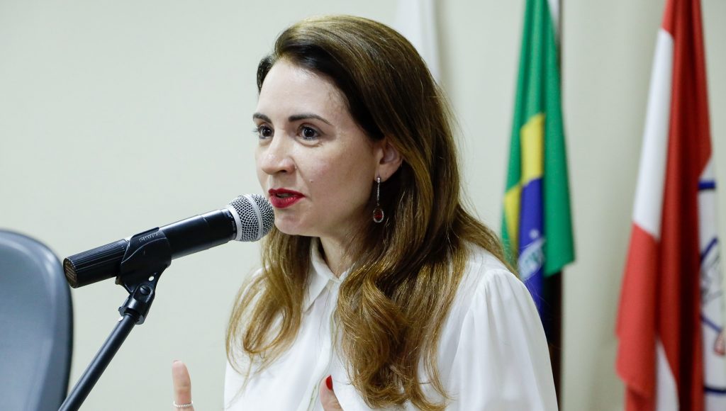 Fotografia de perfil da vice-reitora da UFPA, Loiane Prado Verbicaro, falando ao microfone. Ao fundo, é possível identificar a bandeira do Brasil e a bandeira do Pará.