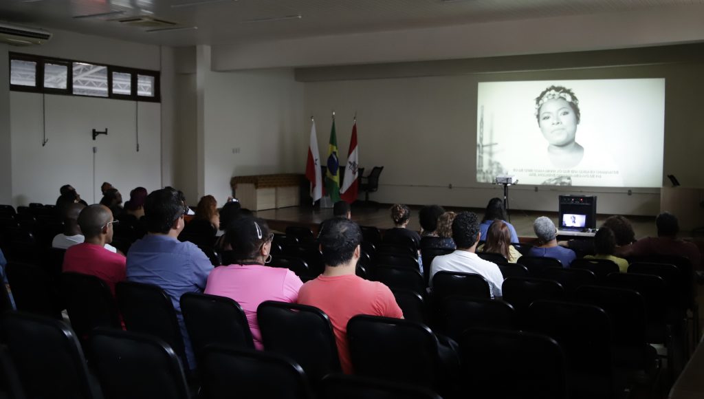 Fotografia registra parte de uma plateia que observam um telão. No telão a imagem de uma mulher jovem.