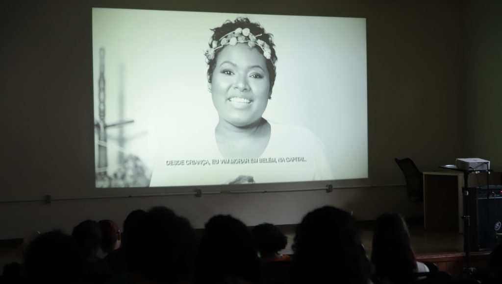 Fotografia registra um ambiente escuro que destaca apenas um grande telão. No telão a imagem de uma mulher jovem, com a legenda "desde criança, eu vim morar em Belém, na capital"