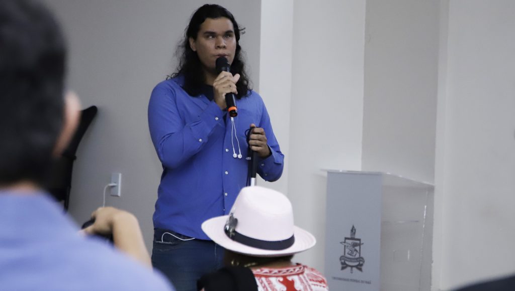 Fotografia registra um jovem falando ao microfone. Ele segura uma bengala na mão esquerda.