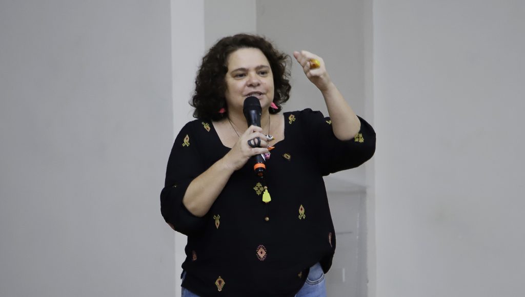 Fotografia registra a professora Milene Lauande falando ao microfone.
