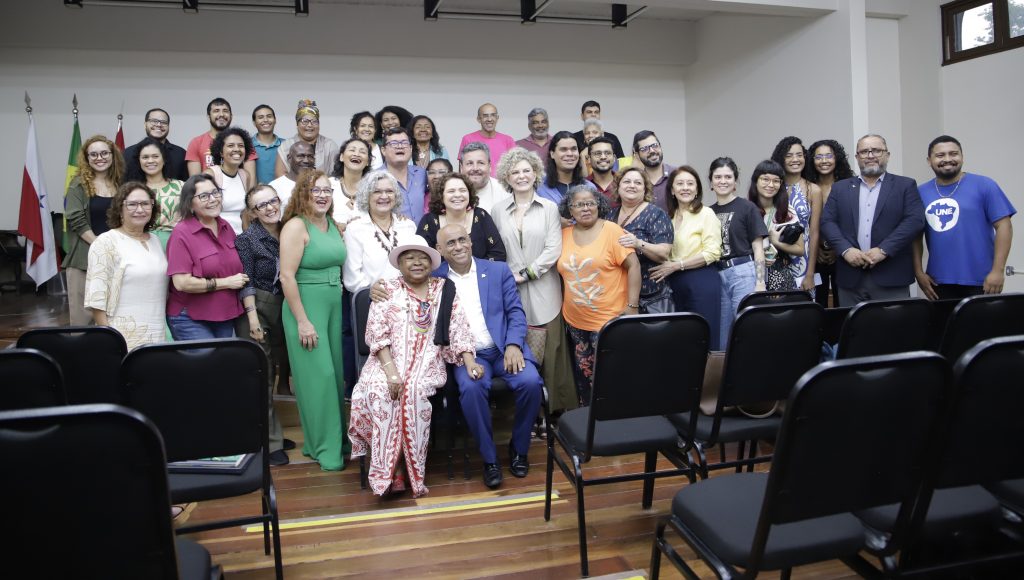 Fotografia posada de um grande grupo de pessoas. Ao centro estão a professora Zélia Amador de Deus e o reitor da UFPA, Gilmar Pereira da Silva. Todos sorriem.