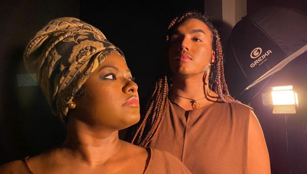 Fotografia posada de duas pessoas, um homem e uma mulher negros. A mulher aparece em primeiro plano, de perfil, usando um turbante. O homem aperece no fundo da foto, usando dread e olhando para a câmera.