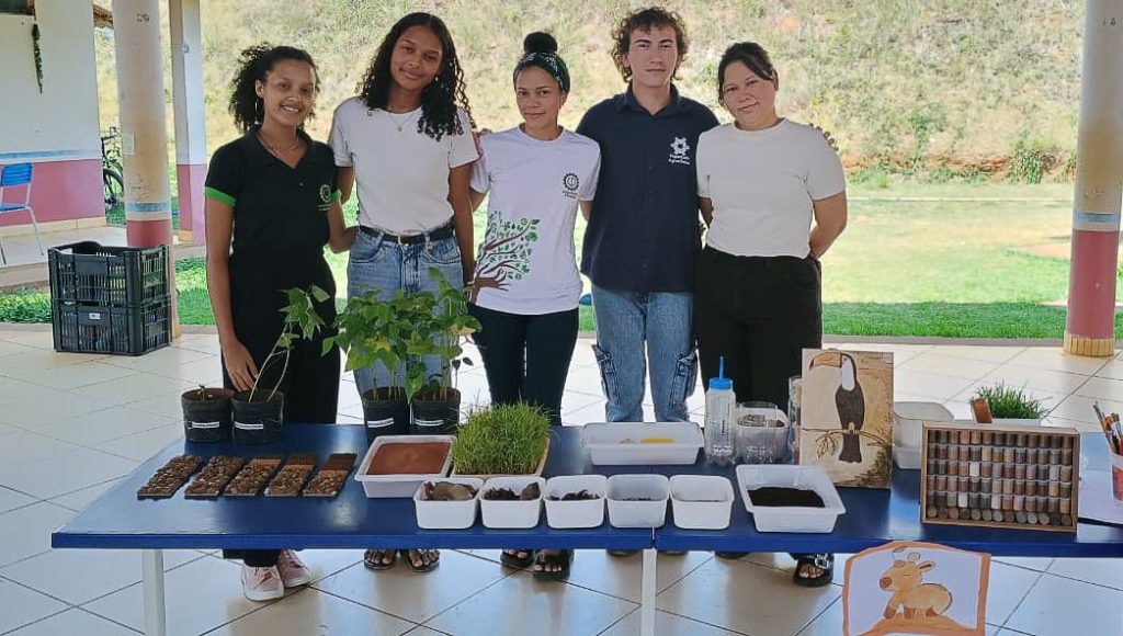 Fotografia posada de cinco pessoas, quatro mulheres e um homem. Eles estão atrás de uma mesa com vários recipientes plásticos com espécies de plantas e terra.