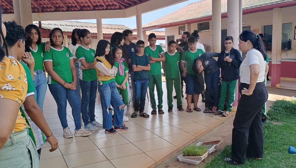 Fotografia registra um grupo de pessoas em um área externa. Uma mulher, próxima a recipientes plásticos com mudas, está de frente para um grupo de jovens uniformizados. Eles obsersavam a mulher.