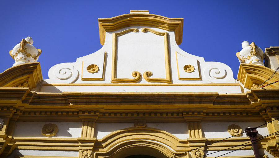 Fotografia de parte da fachada da capela pombo. Na arquitetura, predomina paredes brancas com detalhes em amarelo. Fotografia foi tirada no ângulo de baixo para cima.