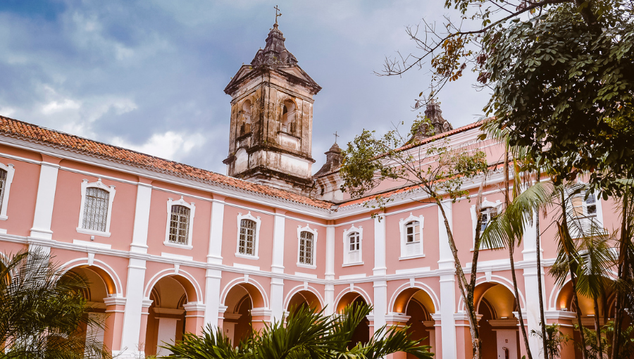 Fotografia mostra parte interma da arquitetura do Convento Mercedários. A estrutura tem predominância da cor rosa com detalhes em branco. Ao fundo, tem-se a torre de uma igreja ainda não restaurada.
