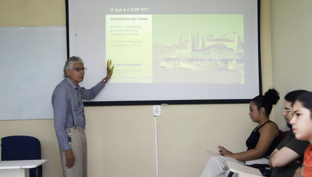 Fotografia registra um homem apontando para uma projeção em um quadro branco. A projeção destaca a seguinte informação: O que é a COP 30?