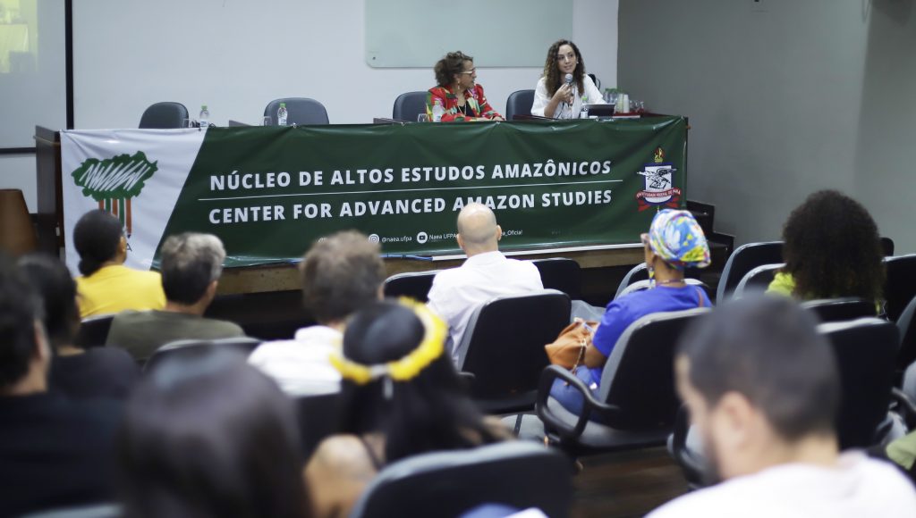 Fotografia registra a mesa de um evento. A mesa está coberta por uma faixa: NÚCLEO DE ALTOS ESTUDOS AMAZÔNICOS. Na mesa, há duas mulheres sentadas. Uma delas fala ao microfone.