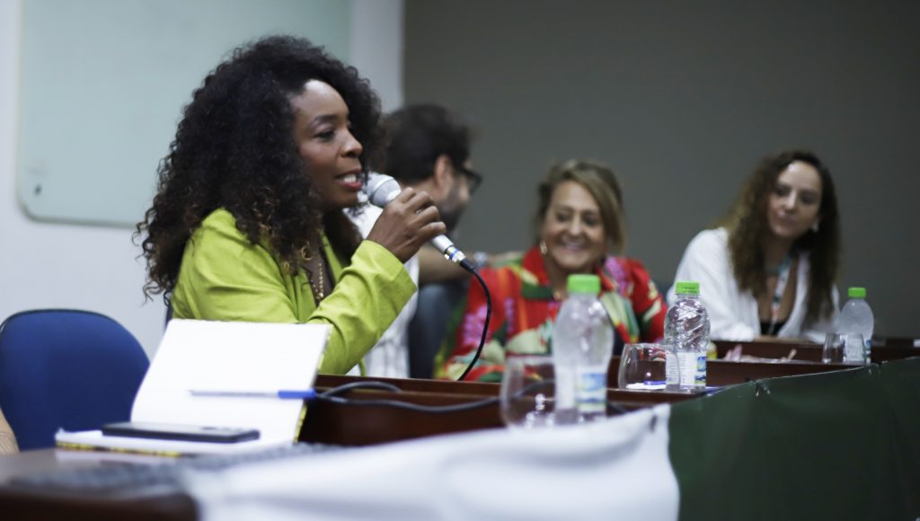 Fotografia registra uma mulher falando ao microfone, enquanto é observada por outras pessoas da mesa.