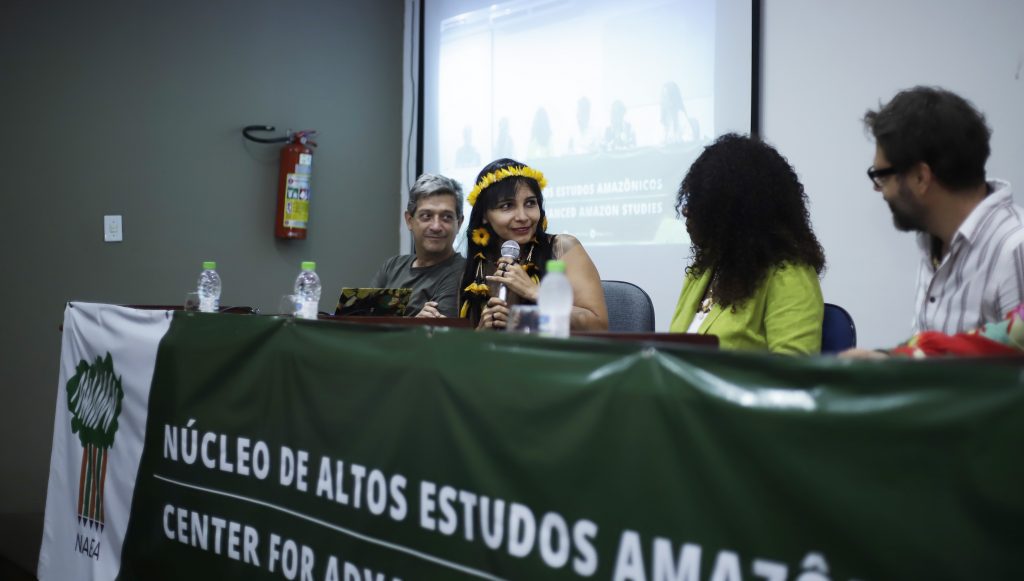 Fotografia registra a mesa de um evento que é formada por quatro pessoas. Um mulher, ao centro, usando um cocar, fala ao microfone.
