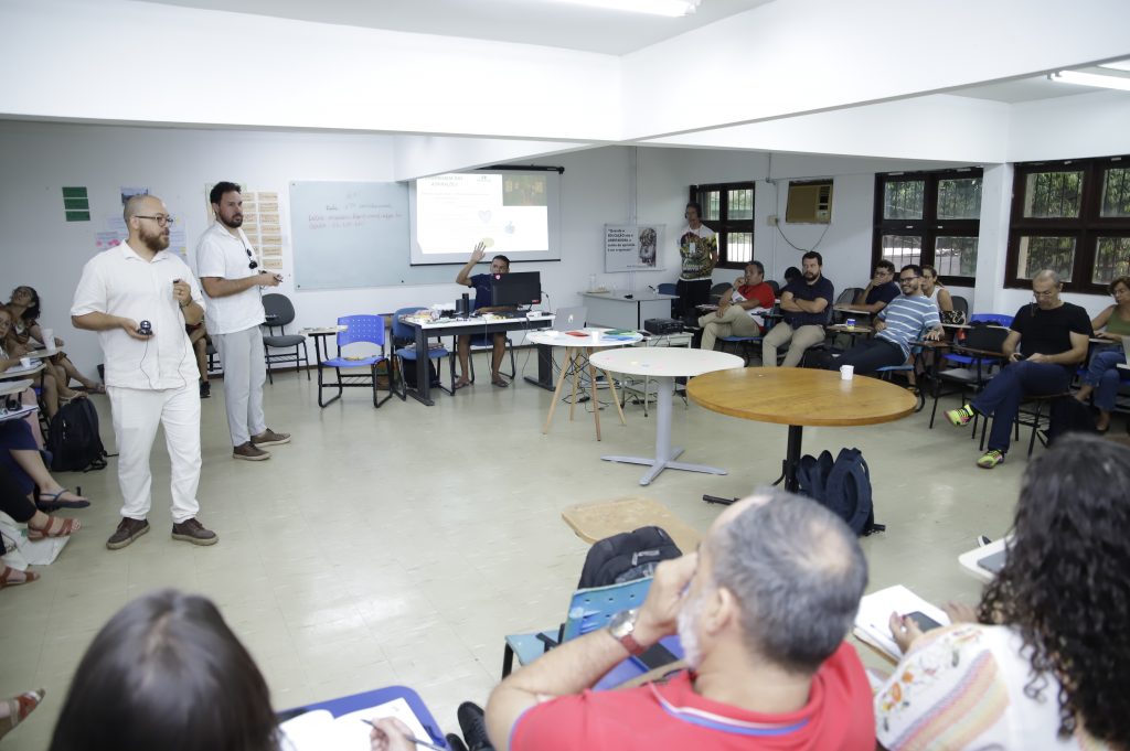 Fotografia mostra uma ampla sala com diversas pessoas sentadas em cadeiras dispostas em uma semi-círculo. Ao centro, dos homens, em pé falam olhando para as pessoas sentadas.