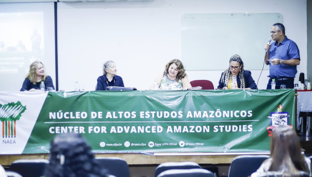 Fotografia registra a mesa de um evento. A mesa está coberta por uma faixa: NÚCLEO DE ALTOS ESTUDOS AMAZÔNICOS. Na mesa, há quatro mulheres sentadas e um homem em pé, falando ao microfone.