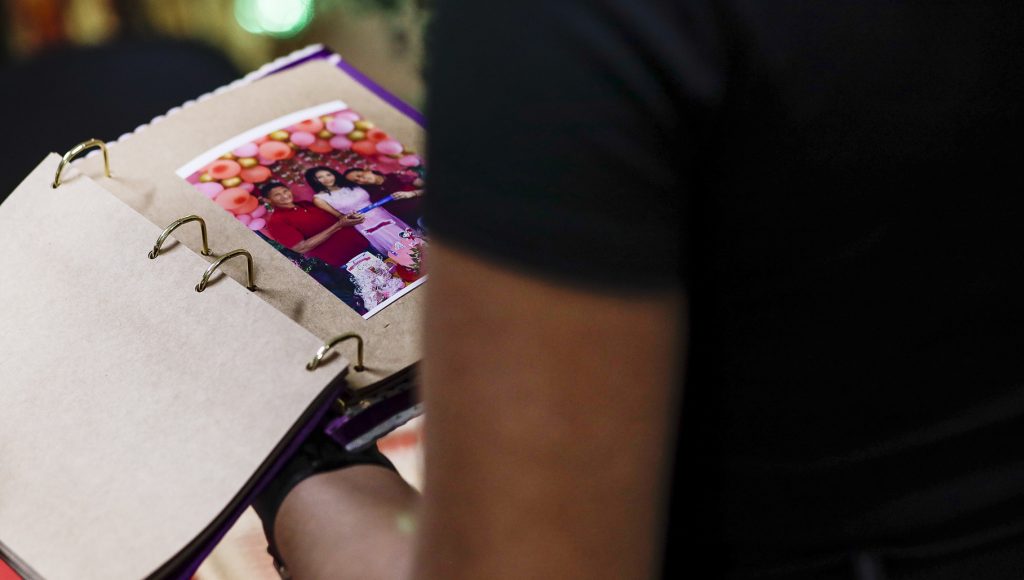 Fotografia registra uma pessoa manuseando um album de fotos. A foto que aparece é de três pessoas posando em um ambiente com balões. uma delas segura um canudo de diploma de formatura.