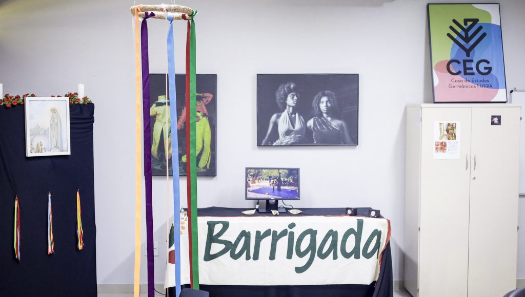 Fotografia registra parte do ambiente da exposição. Há uma placa Barrigada, um quadro da CEG e fitas coloridas penduradas.