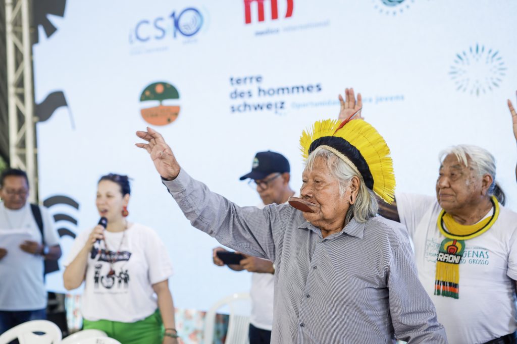 Fotografia mostra Cacique Raoni acenando. Ele está de pé e usar cocar de penas amarelas. Atrás dele há outras pessoas que também acenam.