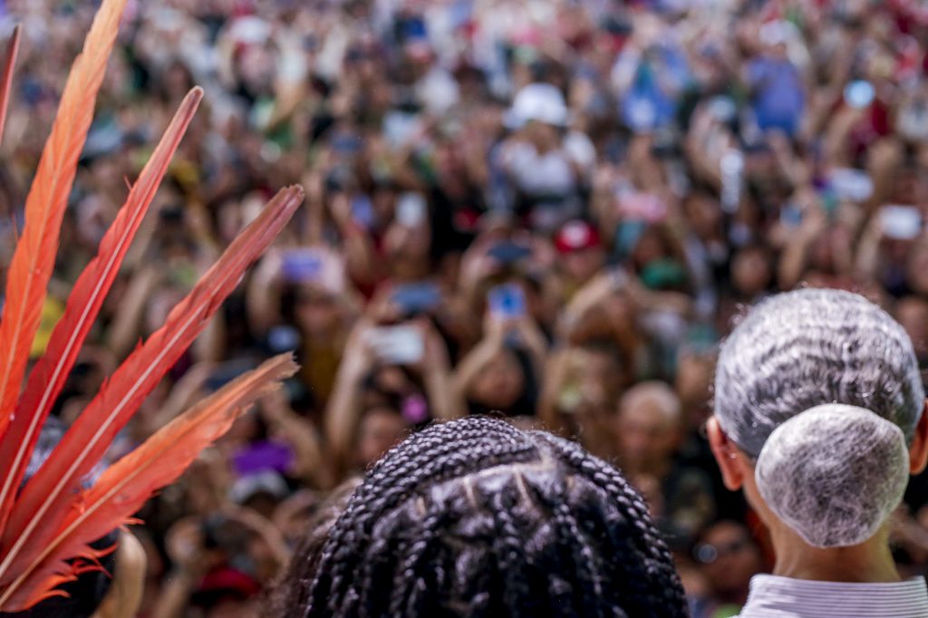 Foto mostra em detalhes parte de um cocar com penas vermelhas, de uma cabeça com tranças e um cabelo grisalho em coque . A imagem é com àngulos das costas para o público que está desfocado na imagem.