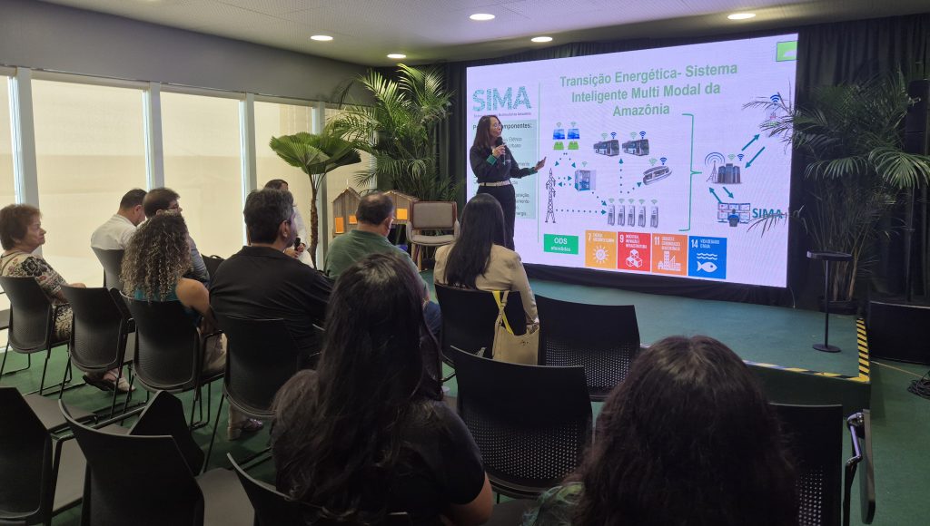 Fotografia registra a professora Emília Tostes, falando ao microfone, durante sua apresentação na Greenzone. Ela é observado por pessoas sentadas na plateia.