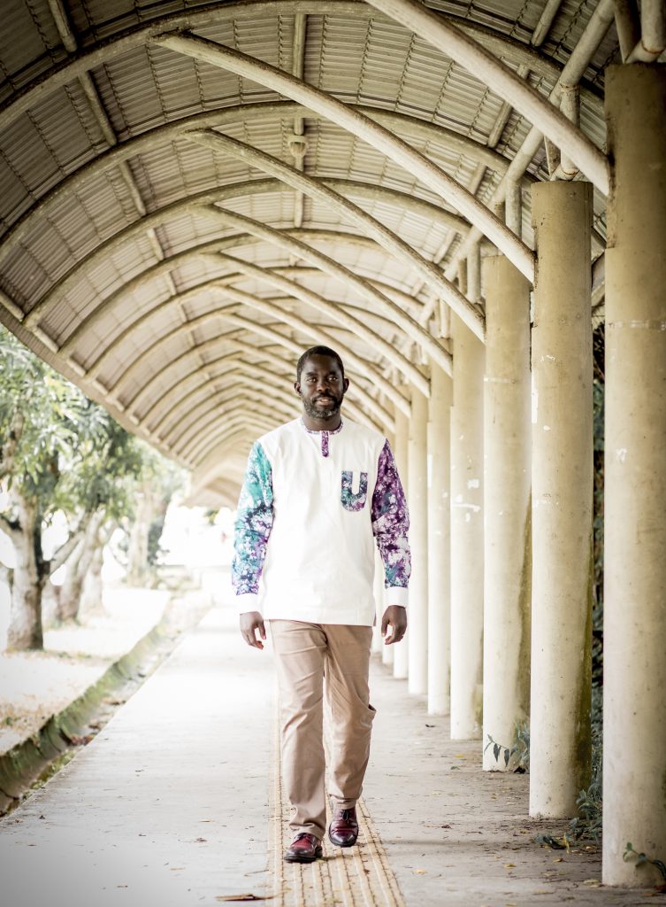 Fotografia posada de Miguel de Barros. Ele caminha por um longo corredor de estrutura branca.