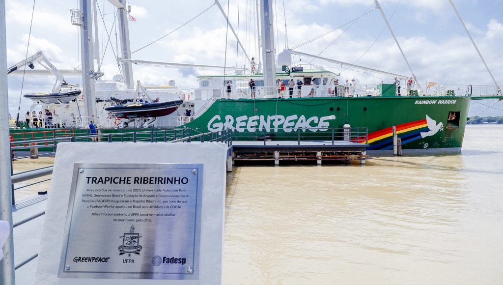 Fotografia registra uma placa de identificação: TRAPICHE RIBEIRINHO, abaixo da informação há o brasão da UFPA e as logomarcas do Greenpeace e da Fadesp. Ao fundo, é possível ver uma grande embarcação na cor verde, identificada com Greenpeace, seguida por um desenho de arco íris e uma pomba branca, com a a descrição "rainbow warrior".