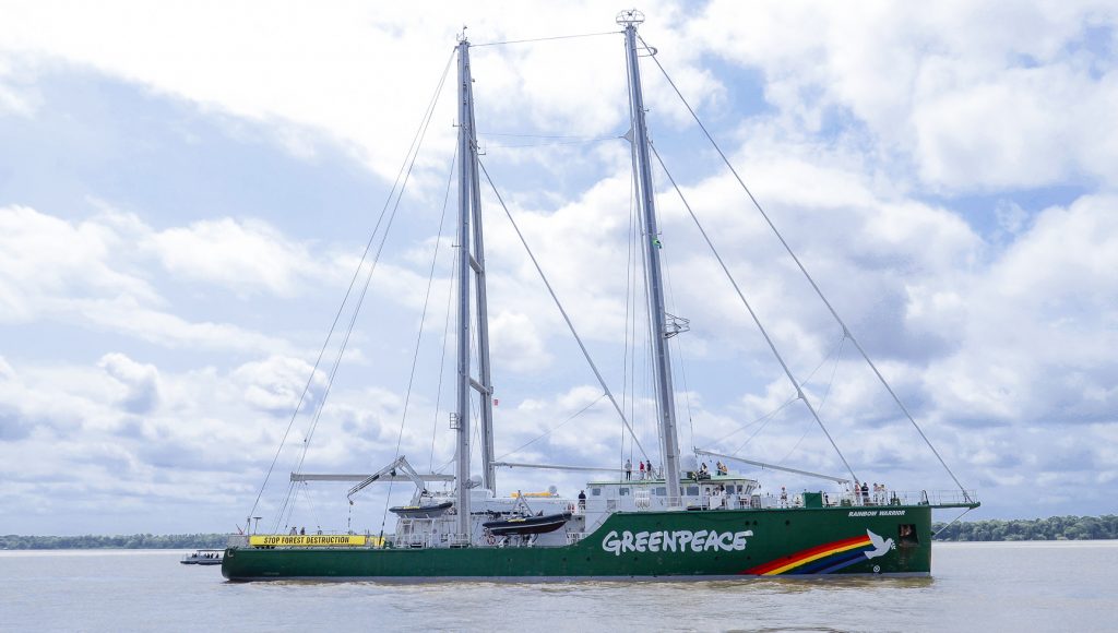 Fotografia de uma grande embarcação na cor verde, identificada com Greenpeace, seguida por um desenho de arco íris e uma pomba branca, com a a descrição "rainbow warrior".