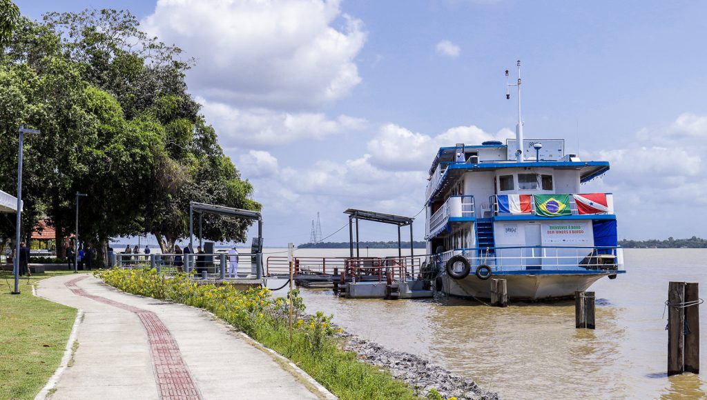 Fotografia registra uma grande embarcação, nas cores branco e azul, atracada em um porto. Na frente da embarcação, estão expostas as bandeiras da França, do Brasil e do Pará.