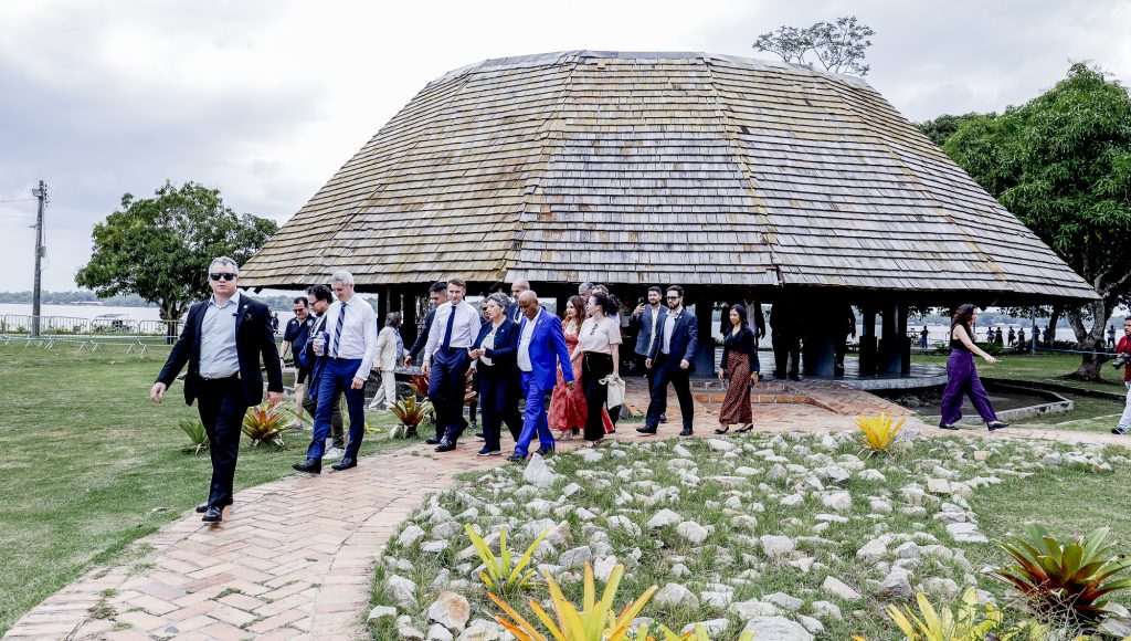 Fotografia registra um grupo de pessoas caminhando por uma passarela. Ao fundo, há uma maloca e árvores. Entre as pessoas, o presidente da França, Emmanuel Macron; o reitor da UFPA, Gilmar Pereira da Silva, e a vice-reitora da UFPA, Loiane Prado Verbicaro.