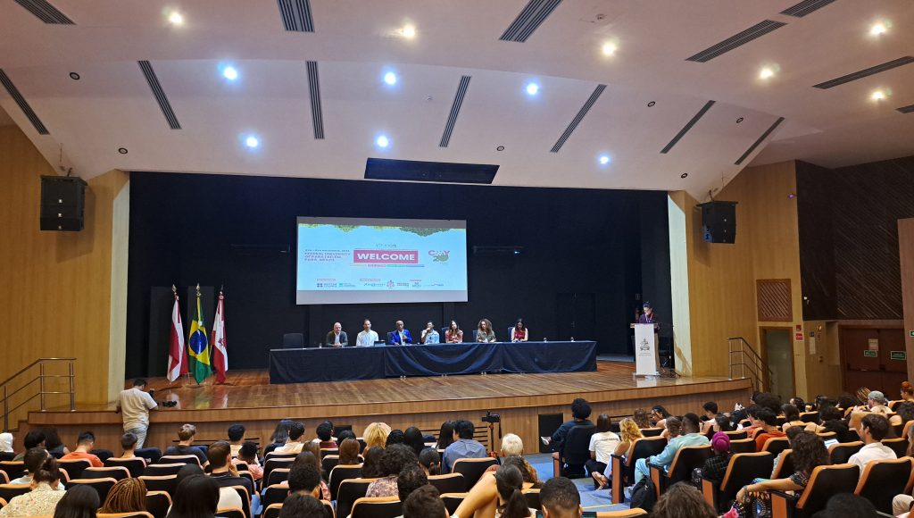Fotografia registra a parte interna de um grande auditório. Ao fundo, no palco, há uma mesa com sete pessoas. Atrás da mesa, um telão exibe a mensagem: WELCOME.