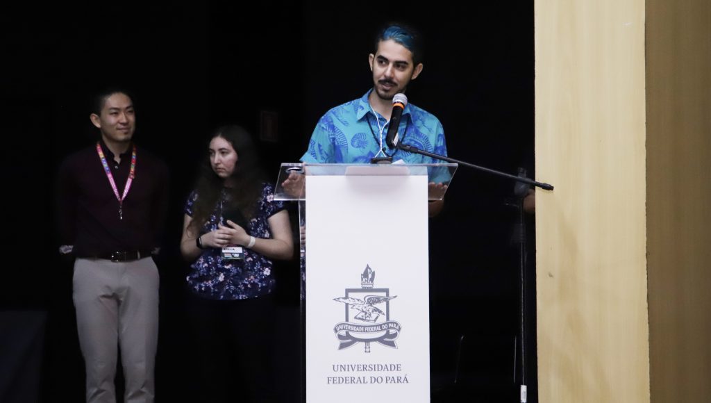 Fotografia registra um homem, jovem, falando ao microfone. O microfone está disposto em um púlpito com o brasão da UFPA.