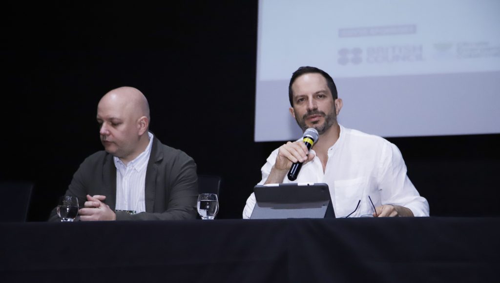 Fotografia registra dois homens sentados na mesa do evento. Um fala ao microfone.