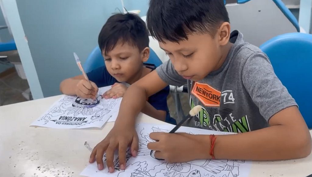 Fotografia registra duas crianças, de cabeças baixas, pintando desenhos em folhas de papel.