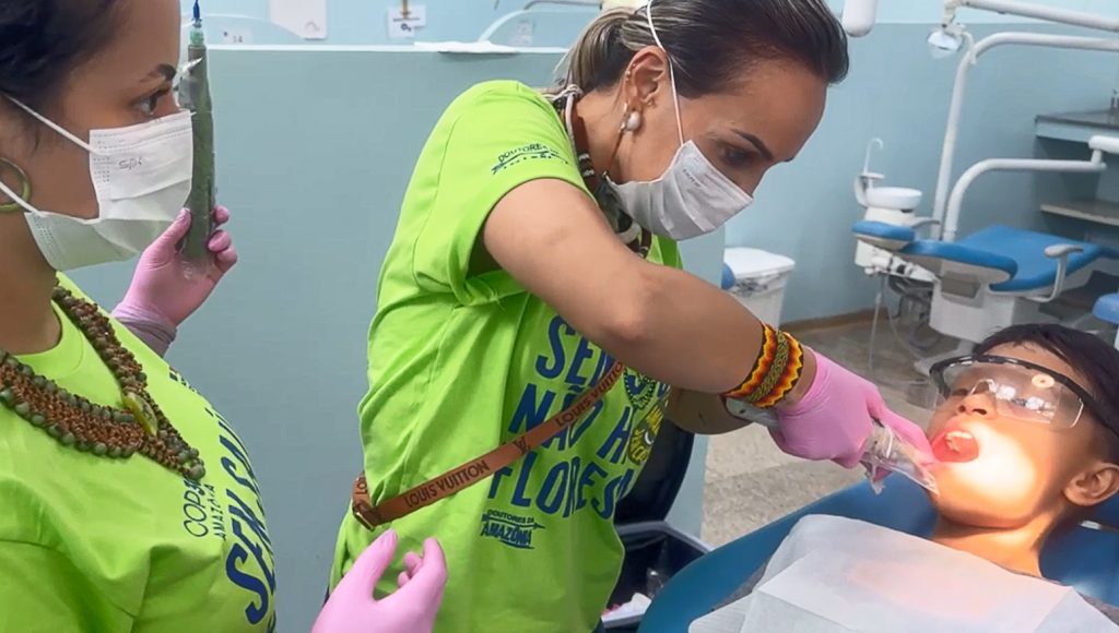 Fotografia registra um atendimento em consultório dentário. Duas mulheres atendem uma criança.