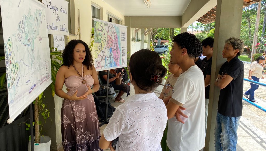 Fotografia registra uma mulher apresentando um painel, em um ambiente aberto, para um grupo de jovens.