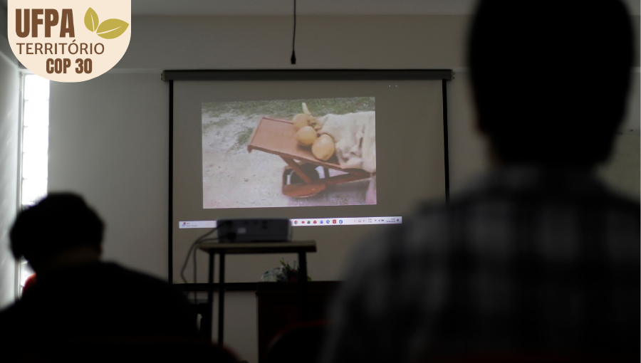 Fotografia registra pessoas de costa atentas a um telão que reproduz um vídeo. Sobre a foto, um selo identifica: UFPA TERRITÓRIO DA COP.