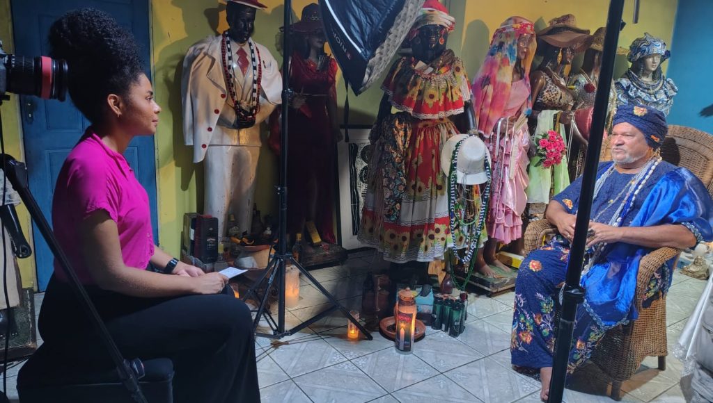 Fotografia registra a estudante Elizabete de Jesus, sentada de frente para um senhor que usa turbante e veste uma grande túnica azul. Ao fundo, algumas imagens de santos típicas de terreiros de religiões de matriz africanas.