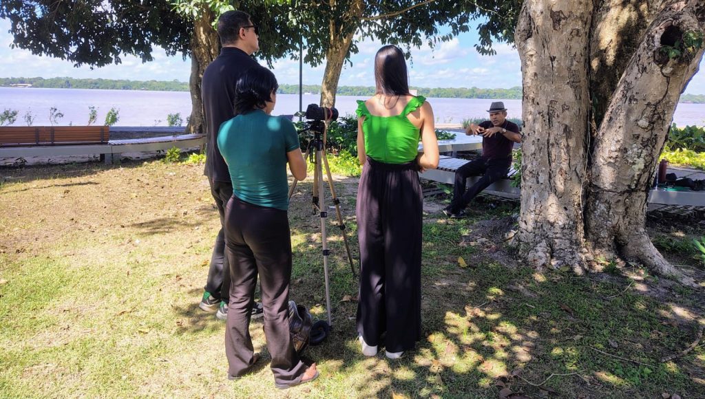 Fotografia registra três pessoas de costa, ao lado de uma câmera que está virada para um homem que está sentado, falando e gesticulando. Eles estão em uma área aberta, com árvores, na beira do rio.