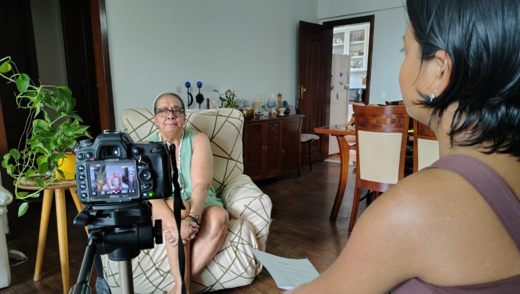 Fotografia registra duas pessoas em uma sala de uma casa. No primeiro plano, é possível identificar uma jovem de costa, ao lado de uma câmera que está focada em uma senhora, que está em segundo plano, sentada em um sofá.