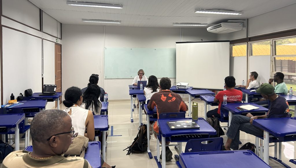 Fotografia de um sala de aula com várias pessoas sentadas em cadeiras azuis, atentas ao fundo da imagem, onde um pessoa está em pé.