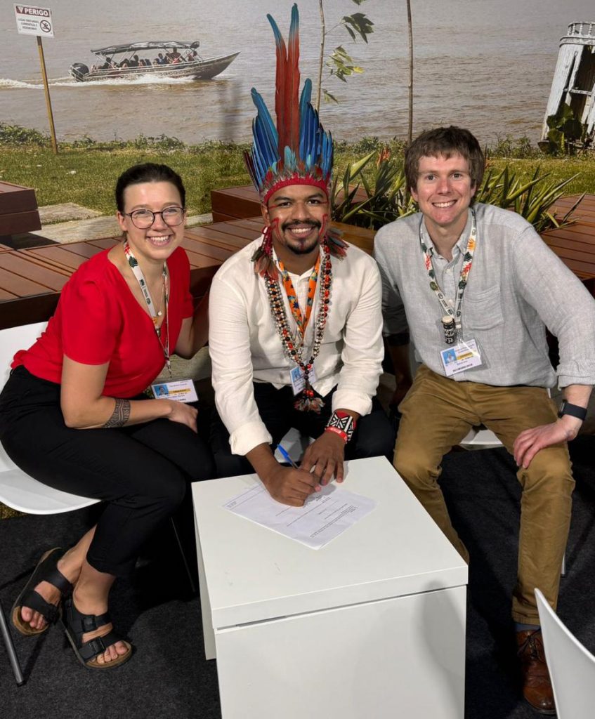 Fotografia posada destaca o estudante indígena Tel Guajajara, acompanhado por outras duas pessoas. Eles estão sentados e Tel assina um papel.