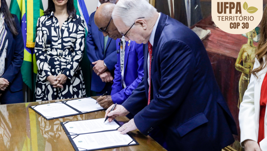 Fotografia registra o reitor da UFPA, Gilmar Pereira da Silva, e o Ministro do Supremo Tribunal Federal, Edson Fachin, assinam documentos disposto sobre uma mesa. Sobre a foto, um selo identifica: UFPA TERRITÓRIO DA COP.