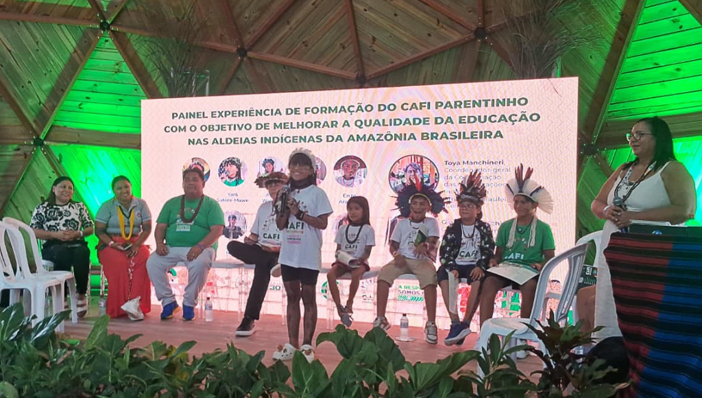 fOTOGRAFIA REGISTRA UM GRUPO DE CRIANÇAS INDÍGENAS. UMA DESSAS CRIANÇAS, UMA MENINA, FALA AO MICROFONE.