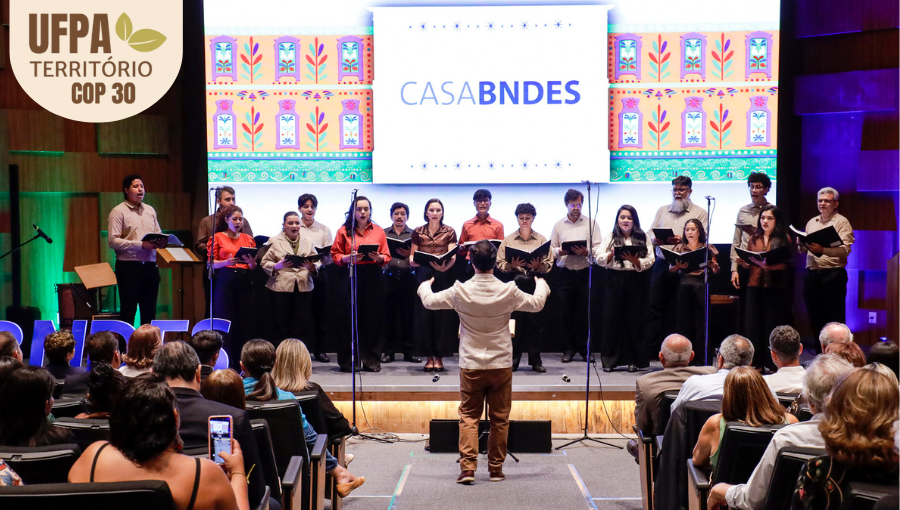 Fotografia registra o palco de um auditório. Há um grupo de canto no palco. De costa para a foto, o maestro rege o grupo. Sobre a foto, um selo identifica: UFPA TERRITÓRIO DA COP.