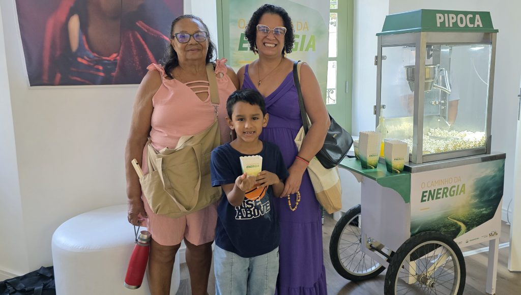 Fotografia posada de duas mulheres adultas e uma criança. Eles estão ao lado de um carrinho de pipoca. A criança, um menino, segura um pacote de pipoca.