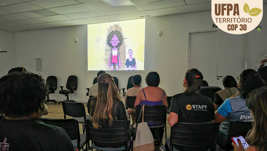 Fotografia registra uma sala de paredes brancas com várias pessoas sentadas de frente para um telão. No telão, é possível ver um imagem de uma boneca e uma tradutora de libras no rodapé do vídeo. Sobre a foto, um selo identifica: UFPA TERRITÓRIO DA COP.