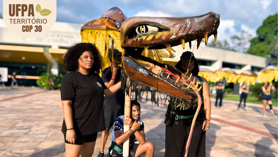 Fotografia registra três pessoas segurando uma grande alegoria que reproduz a cabeça de uma cobra com a língua pra fora. Sobre a foto, um selo identifica: UFPA TERRITÓRIO DA COP.